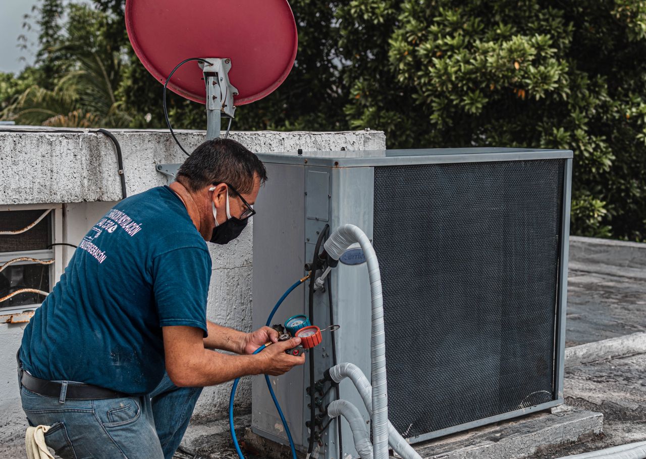 hero-img-02 Technician repairing an air conditioner unit outdoors, wearing a facemask and using a manifold gauge.